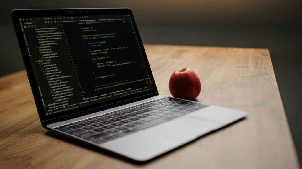 Open laptop displaying code next to a red apple on a wooden desk.
